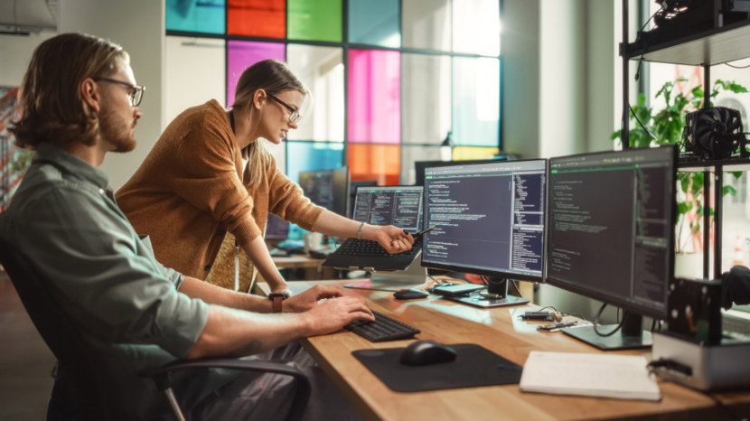 Photo of two people at a workstation desk discussing what is on screen